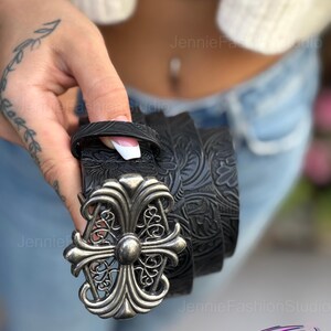 May include: Black leather belt with a decorative silver-colored buckle. The belt features an embossed floral pattern. The buckle has a cross-like design with ornate detailing. The belt is held in a person's hand.