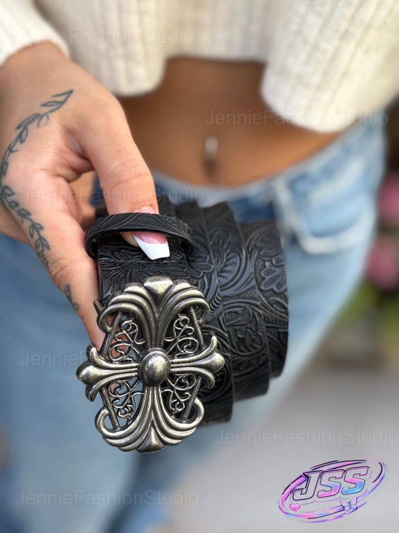 May include: Black leather belt with a decorative silver-colored buckle. The belt features an embossed floral pattern. The buckle has a cross-like design with ornate detailing. The belt is held in a person's hand.