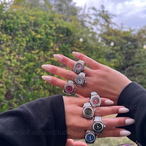 May include: A collection of silver-coloured watch rings with faces in shades of pink, red, white, green, black, and blue. The rings are worn on two hands, highlighting the diverse watch face designs. The backdrop is a blurred view of foliage and the sky.