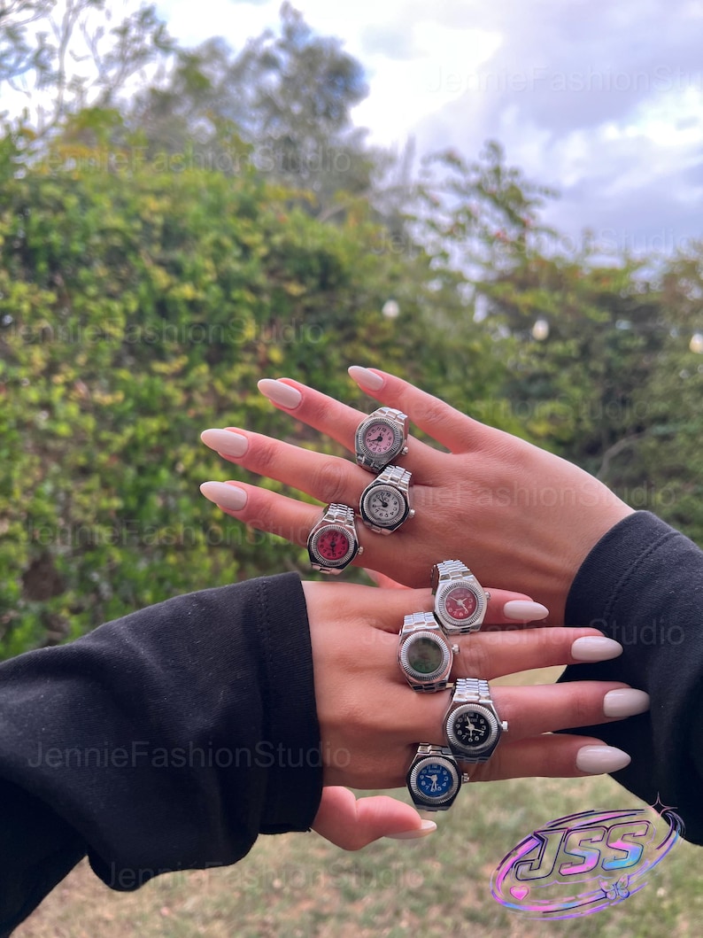 May include: A collection of silver-coloured watch rings with faces in shades of pink, red, white, green, black, and blue. The rings are worn on two hands, highlighting the diverse watch face designs. The backdrop is a blurred view of foliage and the sky.