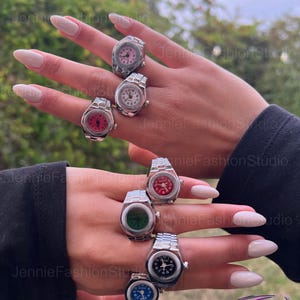 May include: A selection of watch rings featuring silver metal bands and watch faces in a range of colours: pink, white, red, green, black, and blue. The rings are shown on fingers, with a blurred green backdrop.
