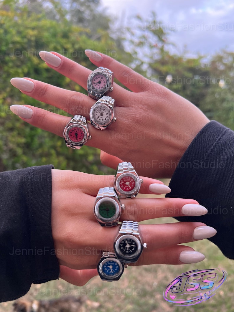 May include: A selection of watch rings featuring silver metal bands and watch faces in a range of colours: pink, white, red, green, black, and blue. The rings are shown on fingers, with a blurred green backdrop.