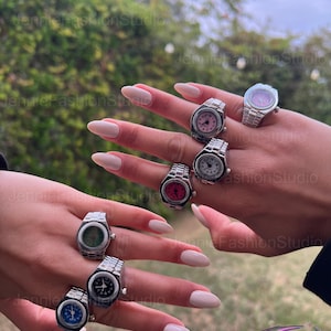 May include: A selection of silver watch rings with different coloured faces, such as pink, red, blue, and green. The rings are shown on a hand with manicured nails, highlighting the jewellery's design. The rings have a watch band style.