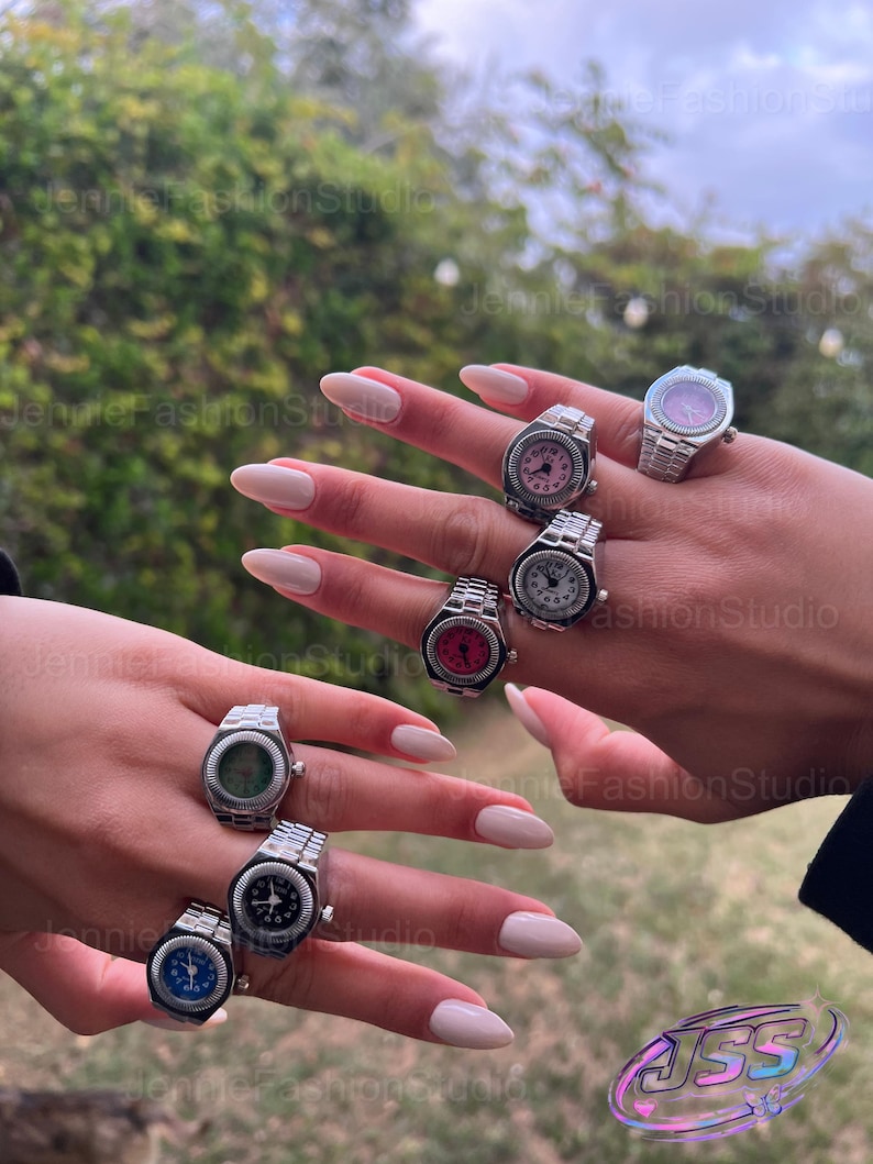 May include: A selection of silver watch rings with different coloured faces, such as pink, red, blue, and green. The rings are shown on a hand with manicured nails, highlighting the jewellery's design. The rings have a watch band style.