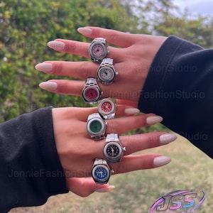 May include: A collection of silver-toned watch rings with various colored faces, including pink, white, red, green, black, and blue. The rings are displayed on a hand with long, manicured nails, showcasing the jewelry's design and detail.