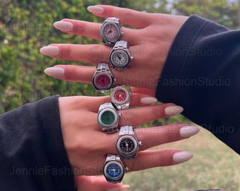 May include: A collection of silver-toned watch rings with various colored faces, including pink, white, red, green, black, and blue. The rings are displayed on a hand with long, manicured nails, showcasing the jewelry's design and detail.