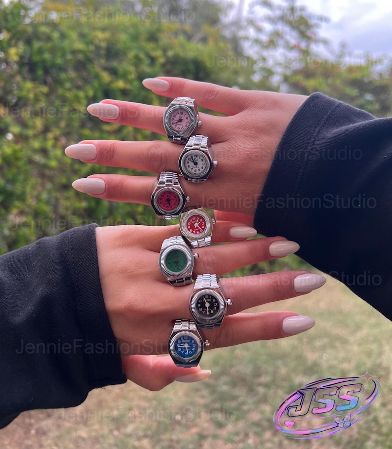 May include: A selection of silver watch rings with different coloured faces, including pink, white, red, green, black, and blue. The rings are displayed on a hand with long, manicured nails, highlighting the jewellery's design and detail.