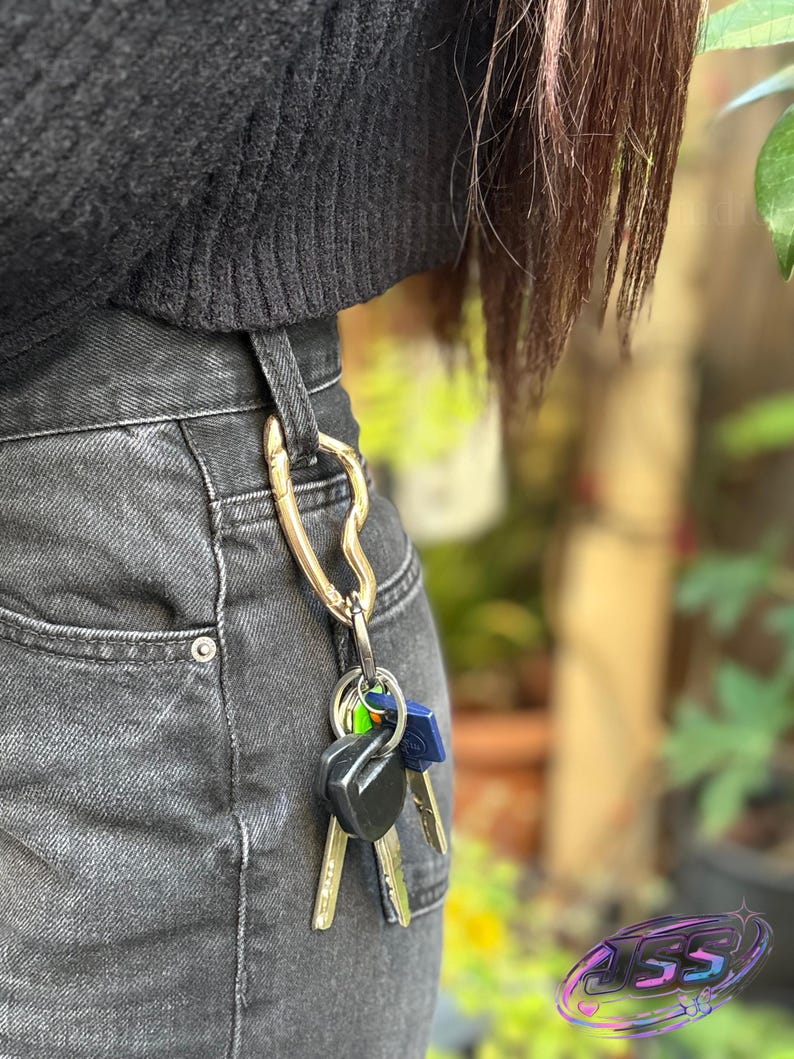 May include: A gold-colored carabiner clip attached to a pair of black denim jeans. Several keys hang from the clip, with a mix of black, blue, and silver key heads. The carabiner is heart-shaped.