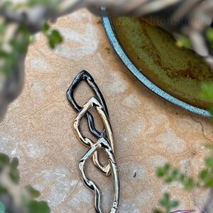 May include: A mixed-metal clasp with a unique, intertwined design. The clasp features sections in silver, gold, and black, creating a striking contrast. The piece is set against a neutral, textured background, with hints of greenery and a decorative bowl in the periphery.
