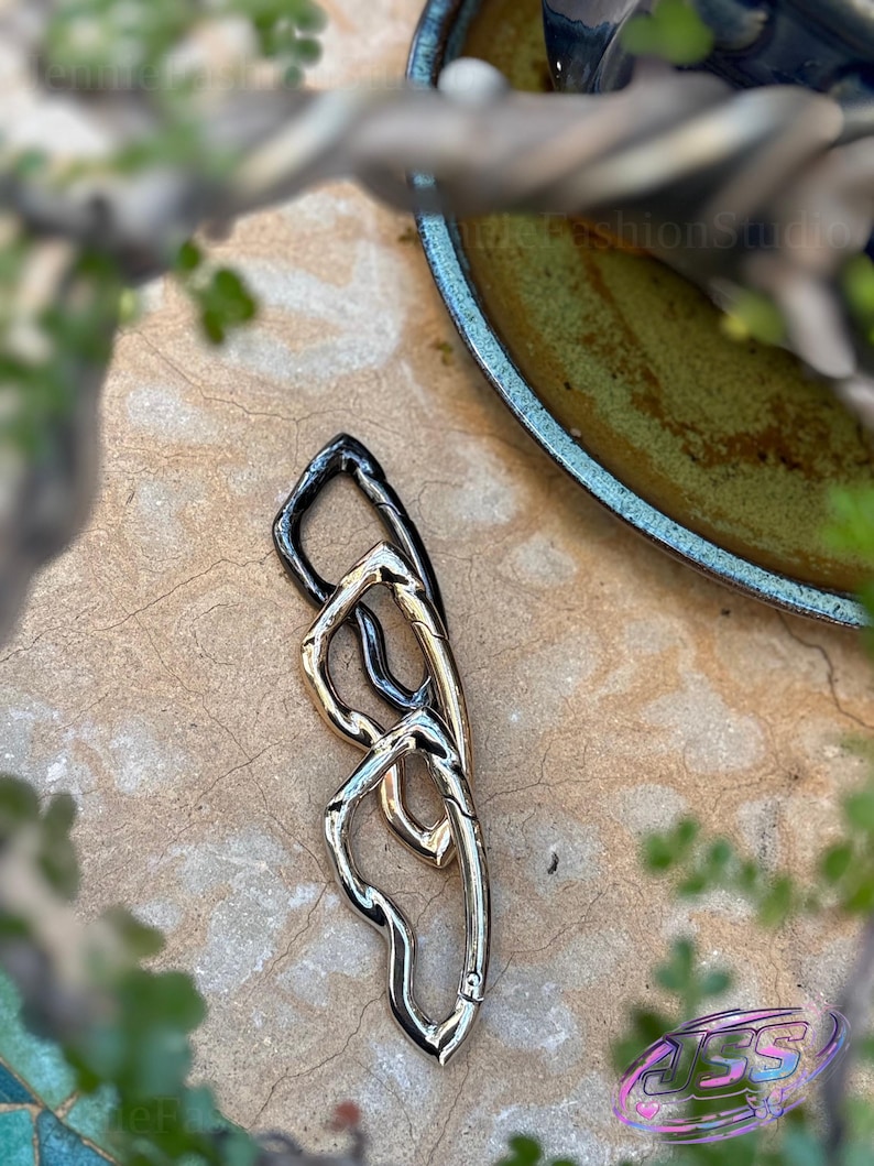 May include: A mixed-metal clasp with a unique, intertwined design. The clasp features sections in silver, gold, and black, creating a striking contrast. The piece is set against a neutral, textured background, with hints of greenery and a decorative bowl in the periphery.