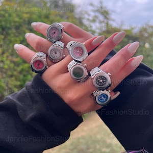 May include: A selection of silver watch rings with faces in shades of pink, white, green, black, and blue. The rings are shown on a hand, highlighting the different watch face designs. These rings are a unique fashion accessory, perfect for adding a touch of style.
