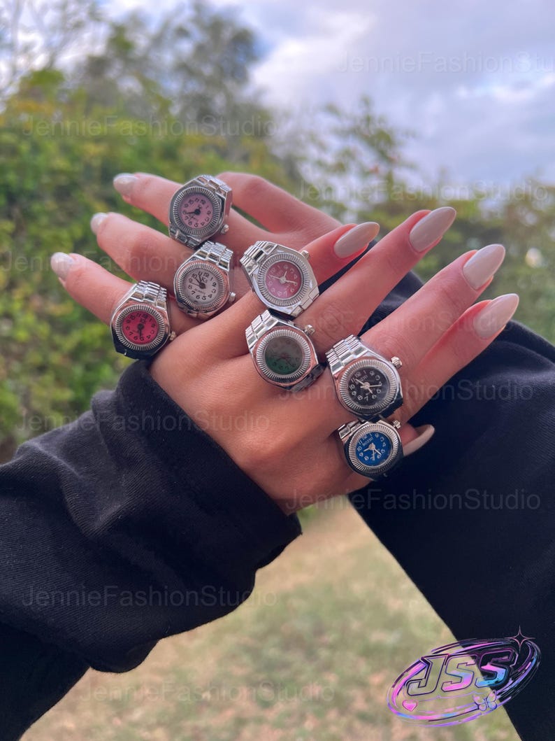 May include: A selection of silver watch rings with faces in shades of pink, white, green, black, and blue. The rings are shown on a hand, highlighting the different watch face designs. These rings are a unique fashion accessory, perfect for adding a touch of style.