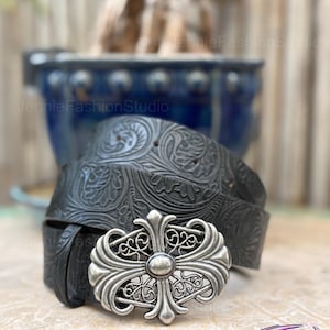 May include: Black leather belt with an ornate silver buckle. The belt features a detailed floral pattern and a decorative silver buckle with a cross design. The belt is a stylish accessory.