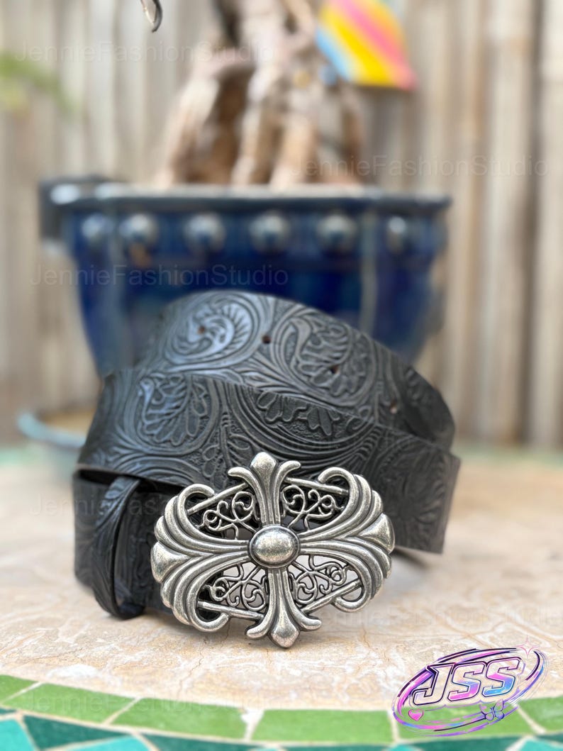 May include: Black leather belt with an ornate silver buckle. The belt features a detailed floral pattern and a decorative silver buckle with a cross design. The belt is a stylish accessory.