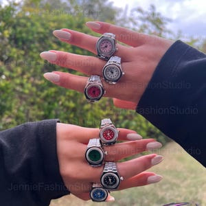 May include: A selection of silver watch rings, each featuring a different coloured watch face: pink, white, red, green, black, and blue. The rings have a traditional watch strap design. The background is a blurred view of green foliage.