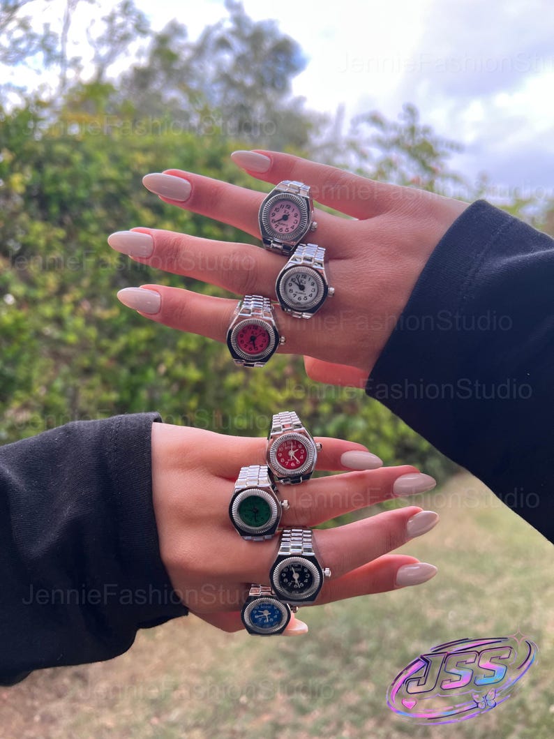 May include: A selection of silver watch rings, each featuring a different coloured watch face: pink, white, red, green, black, and blue. The rings have a traditional watch strap design. The background is a blurred view of green foliage.