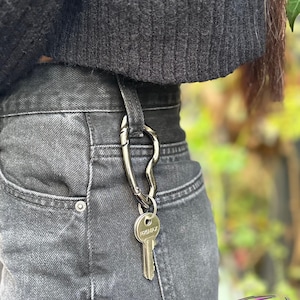 May include: A silver-colored key carabiner attached to a belt loop, holding a key with the text "PREMAX". The carabiner is a unique, curved design. The jeans are a faded black denim.