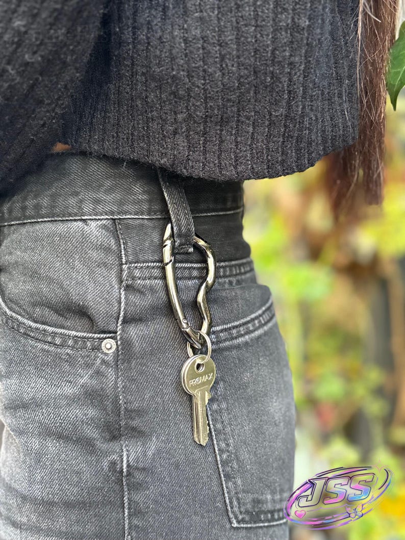 May include: A silver-colored key carabiner attached to a belt loop, holding a key with the text "PREMAX". The carabiner is a unique, curved design. The jeans are a faded black denim.