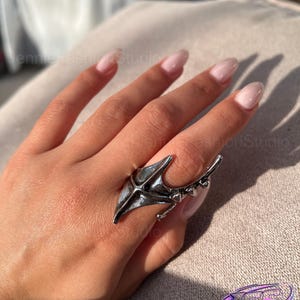 Puede incluir: Un anillo de metal plateado con un diseño ancho en forma de ala que se extiende por el dedo. La superficie tiene un acabado pulido con una sutil oxidación oscura en las hendiduras, y la banda incluye pequeños detalles puntiagudos al costado.