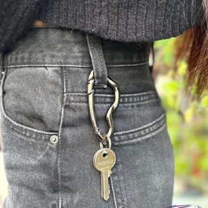 May include: A silver-colored key carabiner clipped to the belt loop of black denim jeans. A single key with the word "PROMAX" is attached to the carabiner. The person is wearing a black sweater.