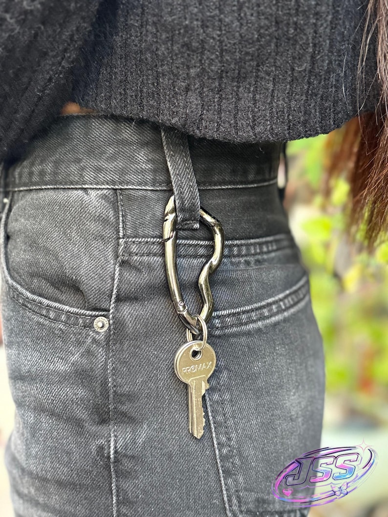 May include: A silver-colored key carabiner clipped to the belt loop of black denim jeans. A single key with the word "PROMAX" is attached to the carabiner. The person is wearing a black sweater.