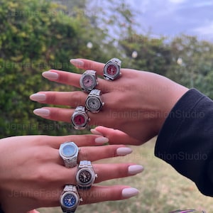 May include: Several silver watch rings with different coloured faces, including red, pink, blue, and black. The rings are shown on two hands against a blurred outdoor backdrop.