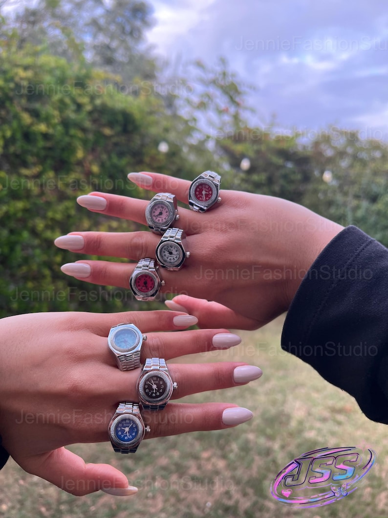 May include: Several silver watch rings with different coloured faces, including red, pink, blue, and black. The rings are shown on two hands against a blurred outdoor backdrop.