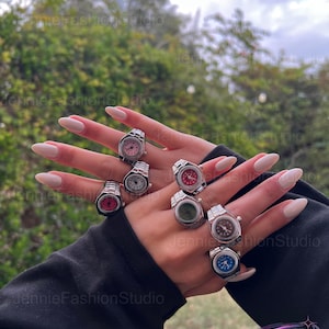 May include: A selection of silver watch rings featuring different coloured faces, such as pink, white, red, green, brown, and blue. The rings are displayed on a hand with manicured nails, set against a natural backdrop.