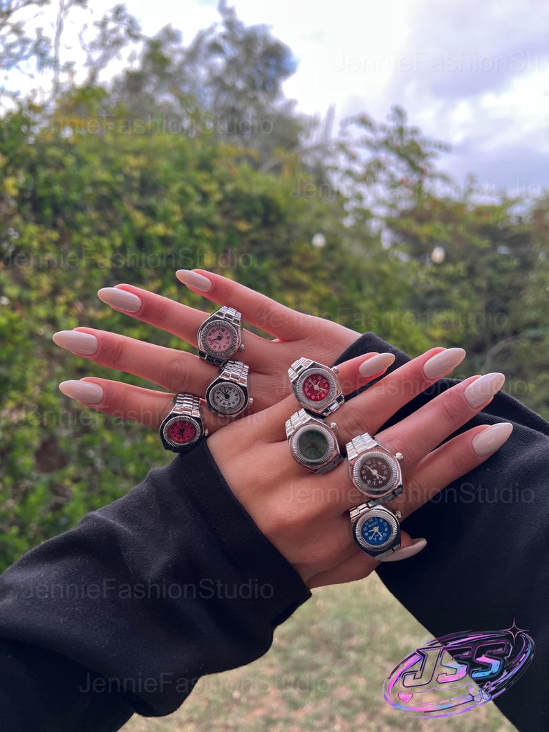 May include: A selection of silver watch rings featuring different coloured faces, such as pink, white, red, green, brown, and blue. The rings are displayed on a hand with manicured nails, set against a natural backdrop.