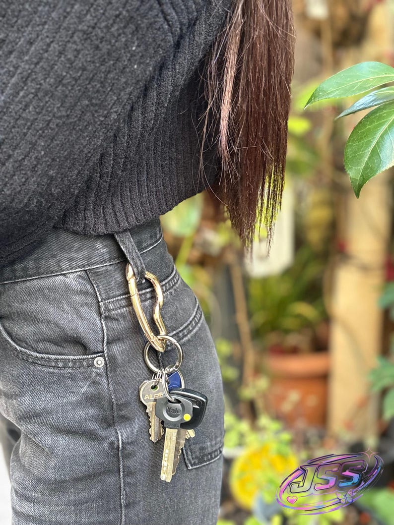 May include: A gold-colored keychain with multiple keys attached to a pair of black denim jeans. The keychain is clipped to the belt loop. The keys include a Yale key and other standard keys.