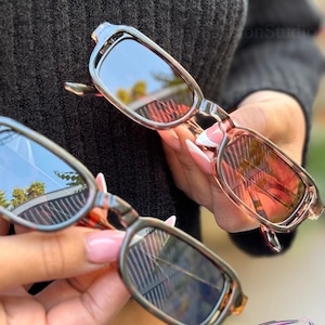 May include: Two pairs of sunglasses with rectangular frames. One pair has a clear frame with pink-tinted lenses, and the other has a tortoiseshell frame with mirrored lenses. The sunglasses are held up against a dark background.