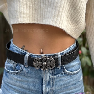 May include: Black leather belt with a silver-colored ornate buckle. The belt is worn with blue denim jeans and a cream-colored sweater. The buckle has a cross-like design. The belt has a floral pattern.