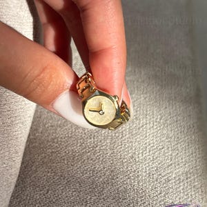 May include: A gold-toned watch ring with a cream-colored face and a small diamond accent. The ring features a rectangular watch face and a linked band. The watch ring is held up against a neutral background.