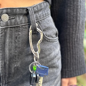May include: A silver-colored key holder with a carabiner clip attached to a pair of black denim jeans. Several keys of different shapes and colors hang from the key ring. The jeans have a button closure and belt loops.