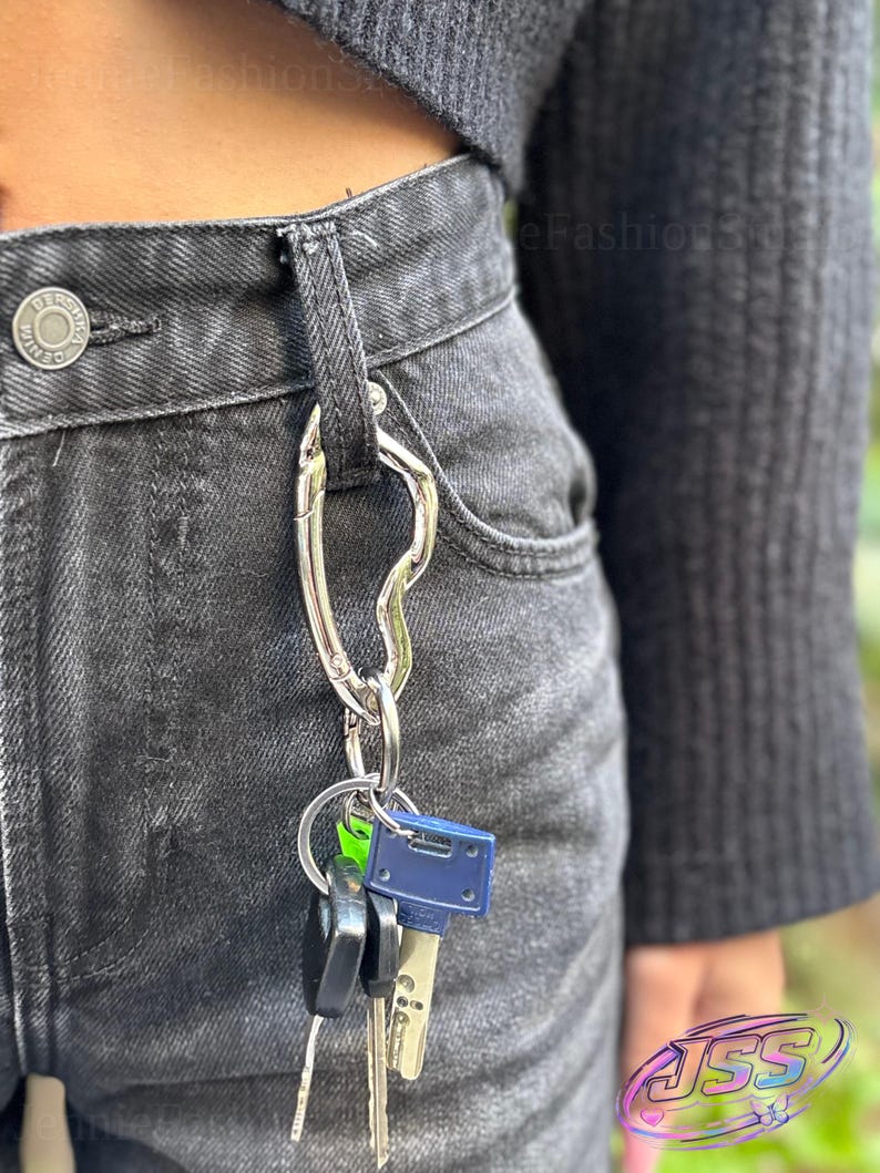 May include: A silver-colored key holder with a carabiner clip attached to a pair of black denim jeans. Several keys of different shapes and colors hang from the key ring. The jeans have a button closure and belt loops.