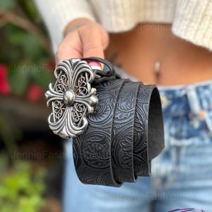 May include: Black leather belt with a silver-colored ornate cross buckle. The belt features a detailed embossed floral pattern. The buckle has a central stud and decorative filigree. A stylish accessory.