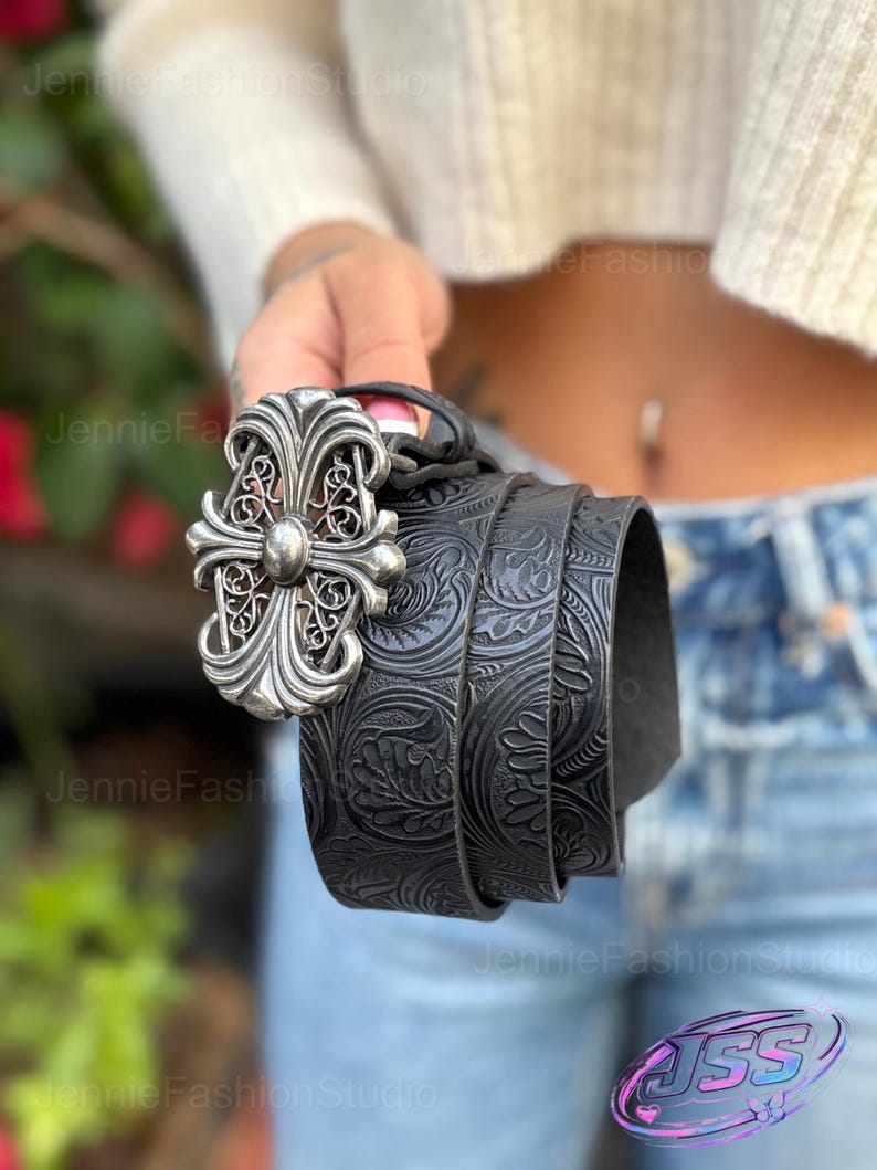 May include: Black leather belt with a silver-colored ornate cross buckle. The belt features a detailed embossed floral pattern. The buckle has a central stud and decorative filigree. A stylish accessory.