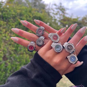 May include: A selection of silver watch rings with different coloured faces, including pink, red, blue, and black. The rings are shown on a hand with long, manicured nails. The background is blurred with green foliage.