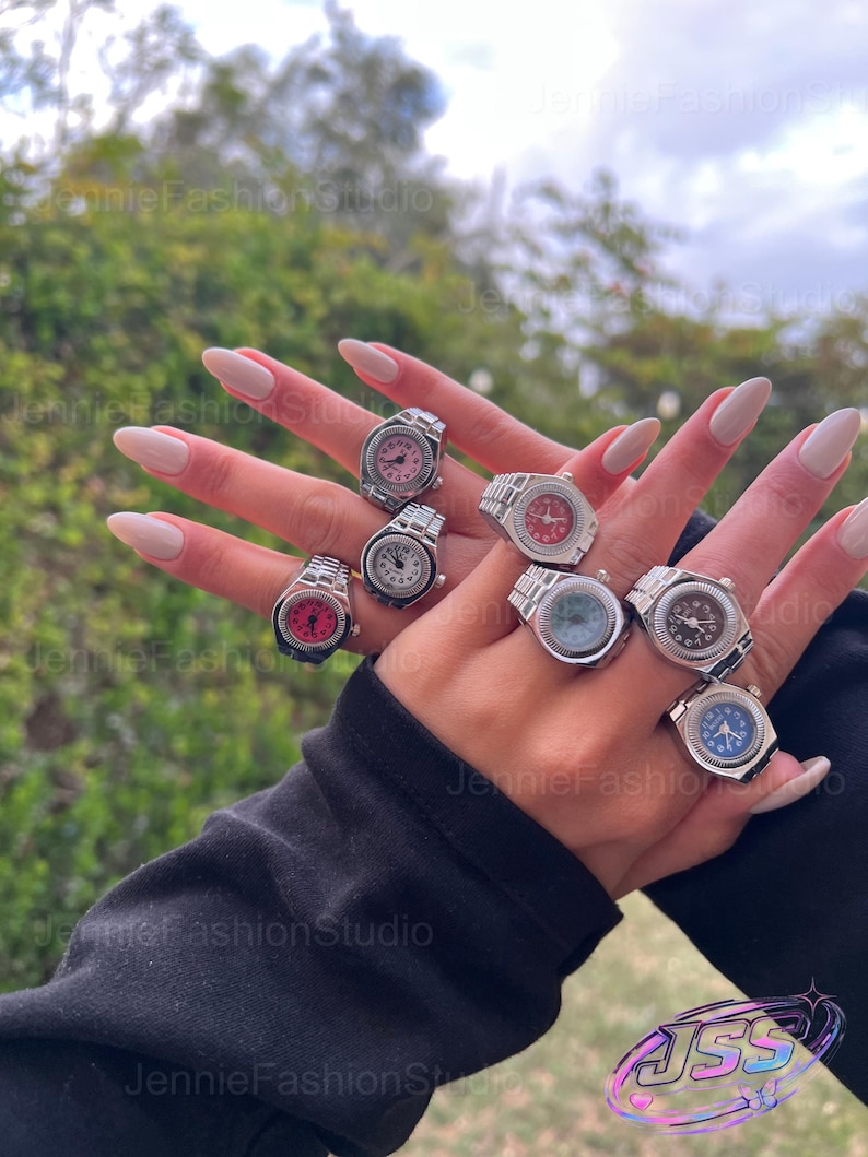 May include: A selection of silver watch rings with different coloured faces, including pink, red, blue, and black. The rings are shown on a hand with long, manicured nails. The background is blurred with green foliage.