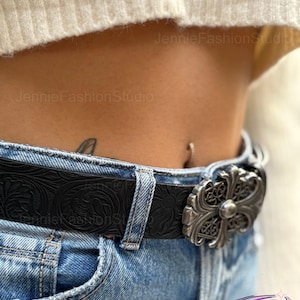 May include: Black leather belt with embossed floral pattern and a silver-colored ornate buckle. The belt is worn with blue jeans. The buckle has a cross-like design.