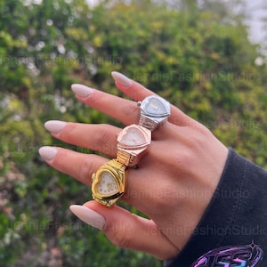 Peut inclure: Trois bagues-montres en forme de cœur, argentées, or rose et dorées. Les montres ont un petit cadran et sont empilées sur un doigt. Le fond est un feuillage vert flou.