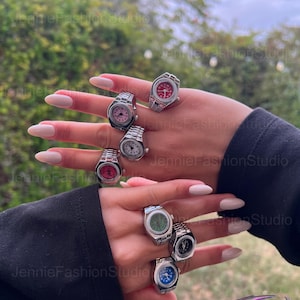 May include: A selection of silver watch rings with faces in shades of pink, red, green, and blue. The rings are shown on a hand with long, manicured nails. The backdrop is a blurred view of green leaves and foliage.