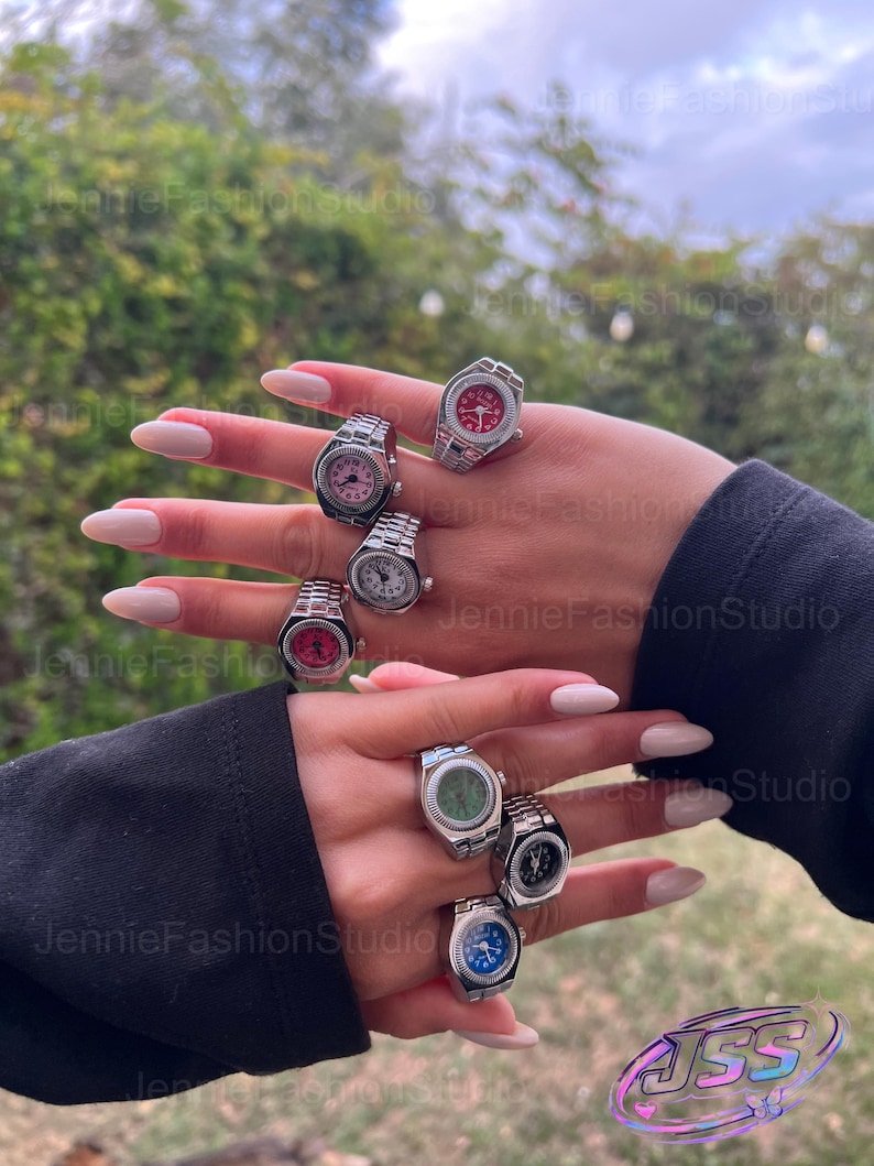 May include: A selection of silver watch rings with faces in shades of pink, red, green, and blue. The rings are shown on a hand with long, manicured nails. The backdrop is a blurred view of green leaves and foliage.