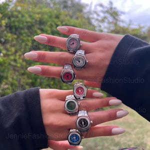 May include: Several silver watch rings with different coloured faces, including pink, white, red, green, black, and blue. The rings are shown on a hand with manicured nails. The background is a blurred view of greenery and sky.