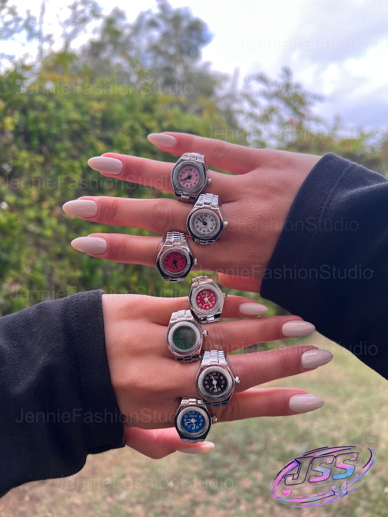 May include: Several silver watch rings with different coloured faces, including pink, white, red, green, black, and blue. The rings are shown on a hand with manicured nails. The background is a blurred view of greenery and sky.