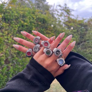 May include: A selection of silver watch rings with different coloured faces, such as pink, red, and blue. The rings are shown on a hand with long, polished nails, set against a background of green foliage and a cloudy sky.