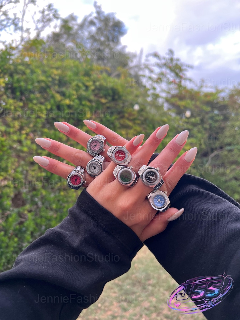 May include: A selection of silver watch rings with different coloured faces, such as pink, red, and blue. The rings are shown on a hand with long, polished nails, set against a background of green foliage and a cloudy sky.