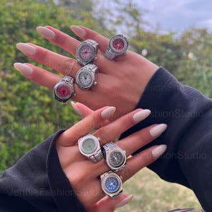 May include: A selection of silver watch rings with different coloured watch faces, including pink, red, white, green, and blue. The rings feature a detailed watch band design. The image is taken outdoors.