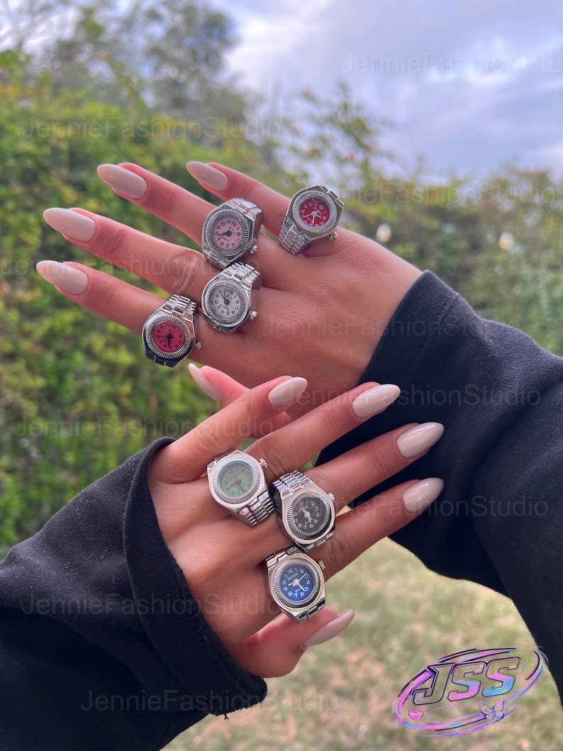 May include: A selection of silver watch rings with different coloured watch faces, including pink, red, white, green, and blue. The rings feature a detailed watch band design. The image is taken outdoors.