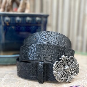 May include: Black leather belt with floral embossed pattern and a silver-colored buckle. The belt is rolled up, showcasing the intricate design. The buckle has a cross-like design.